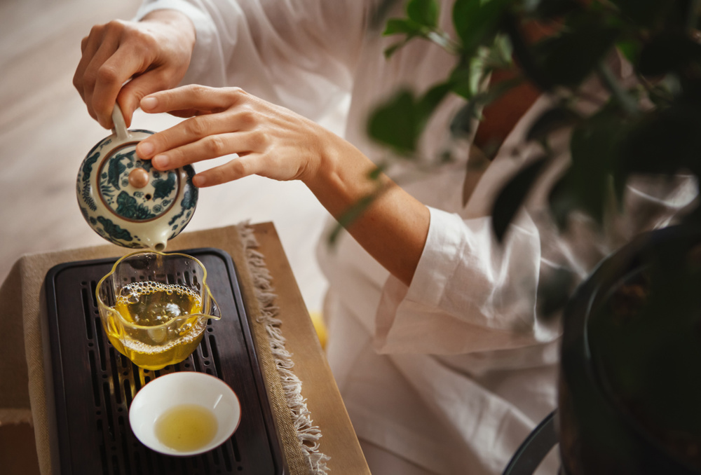 Female hands pouring tea from patterned clay teapot into clear glass pitcher, creating serene tea ritual. Soft light, wooden tea tray and greenery evoke calming mindful Chinese ceremony atmosphere