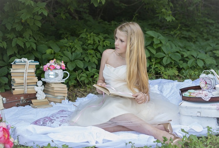 a lady in white dress, sitting and holding a book, with books and a basket of flowers on her side